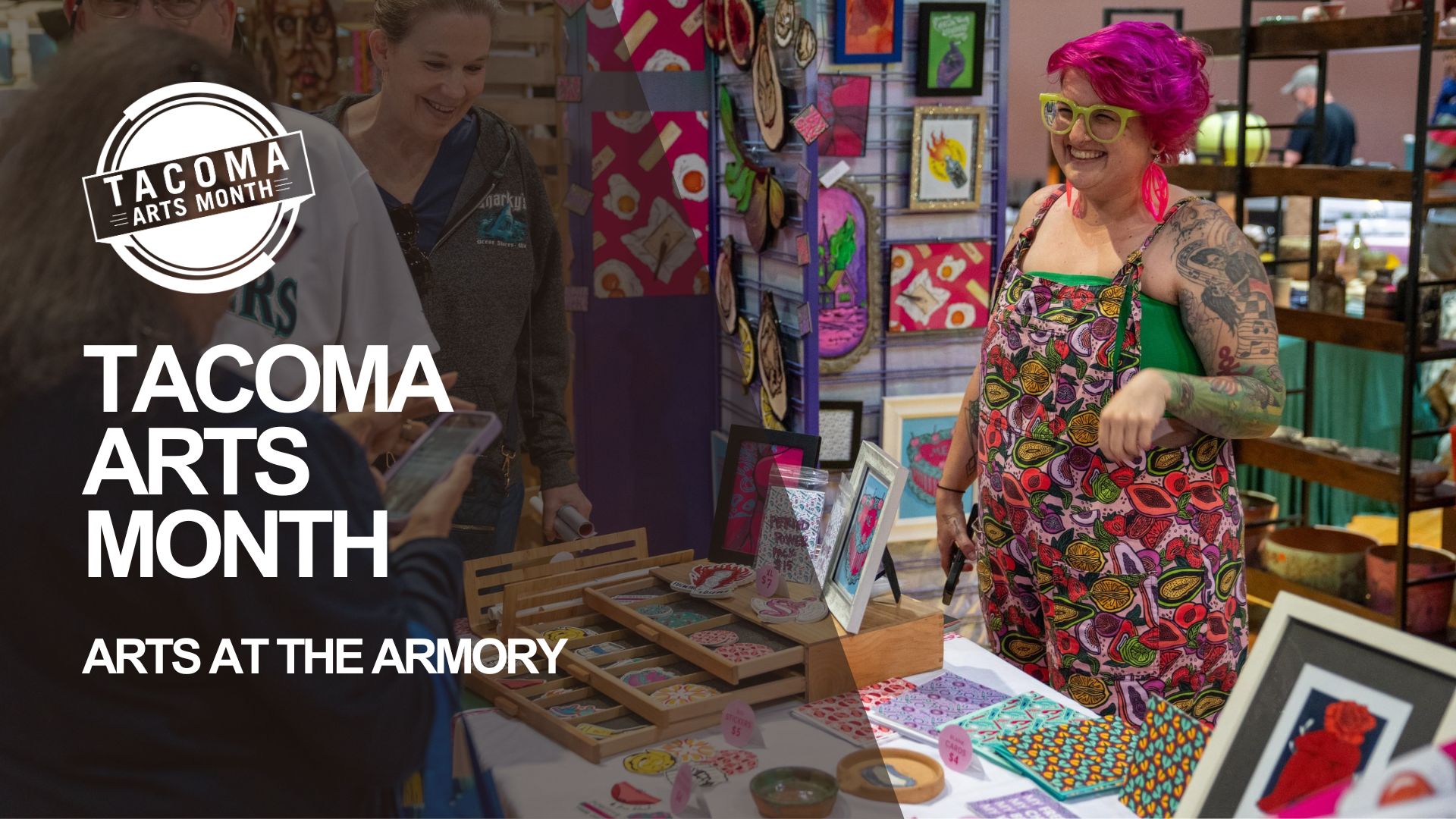 An artist with bright pink hair and glasses wearing a colorful apron smiles while standing at an art booth. Behind them, art is displayed on a panel with a pink background. In front of them, a table displays various art pieces and items as part of Arts at the Armory during Tacoma Arts Month.