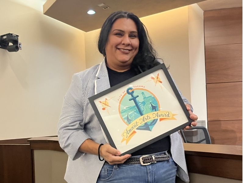 A person holds a framed award that features an anchor and the text "Annual Arts Award" and "City of Tacoma."